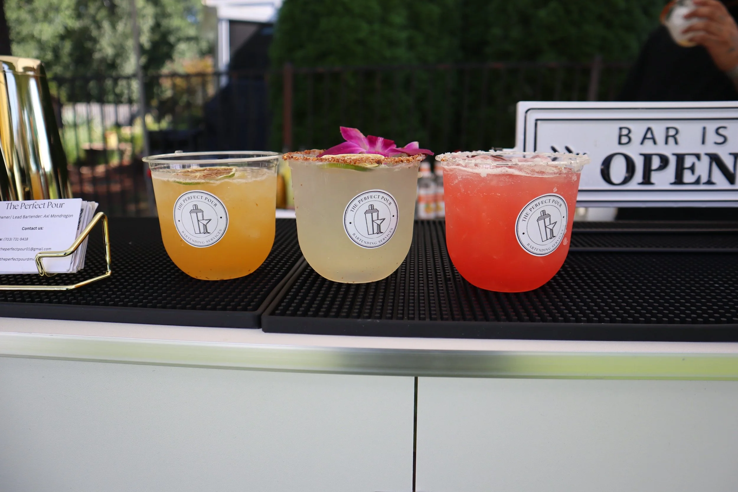 Three colorful cocktails in plastic cups with logo, garnished with lime and flower, placed on a black surface at an outdoor bar with a sign that reads 'Bar is Open' in the background.