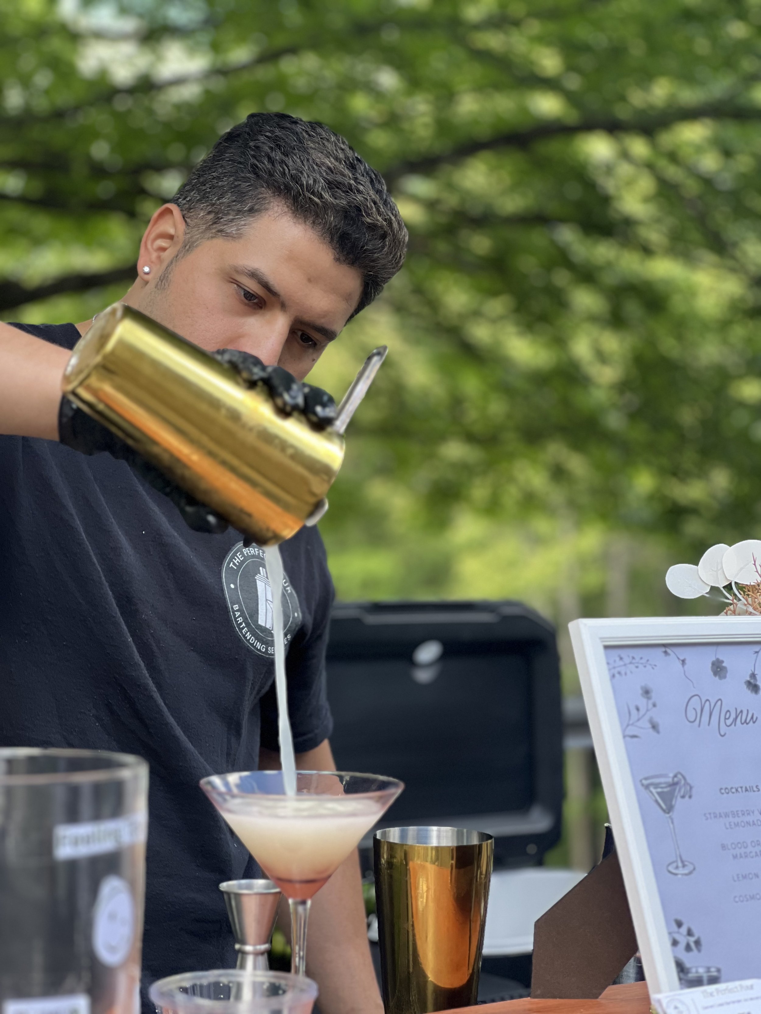 A bartender wearing a black shirt and black gloves is pouring a cocktail from a gold shaker into a martini glass outdoors, with a menu and other bar tools visible on the table, and green trees in the background.