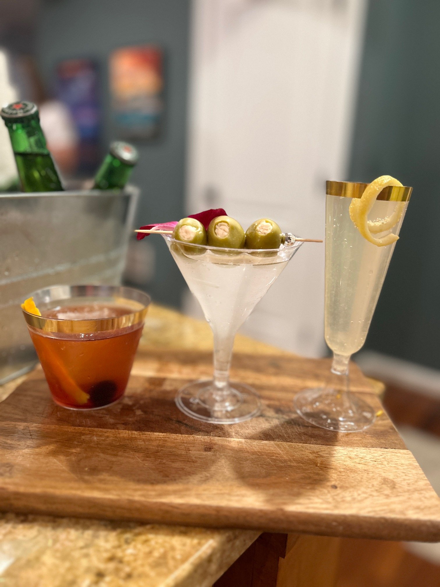 Three cocktails on a wooden tray: a bourbon cocktail with an orange peel and cherry, a martini with stuffed olives, and a champagne flute with a lemon twist. Ice bucket with bottled drinks in the background.