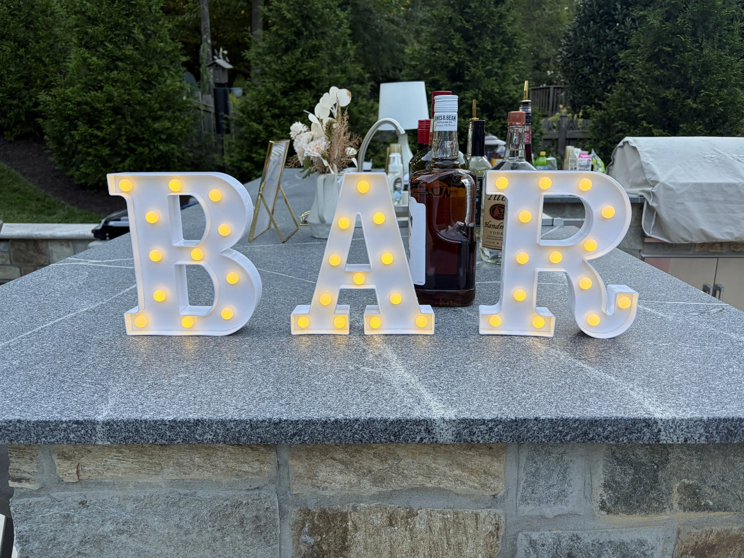Decorative LED letters spelling 'BAR' on a stone ledge, with bottles of alcohol, flowers, and outdoor furniture in the background.