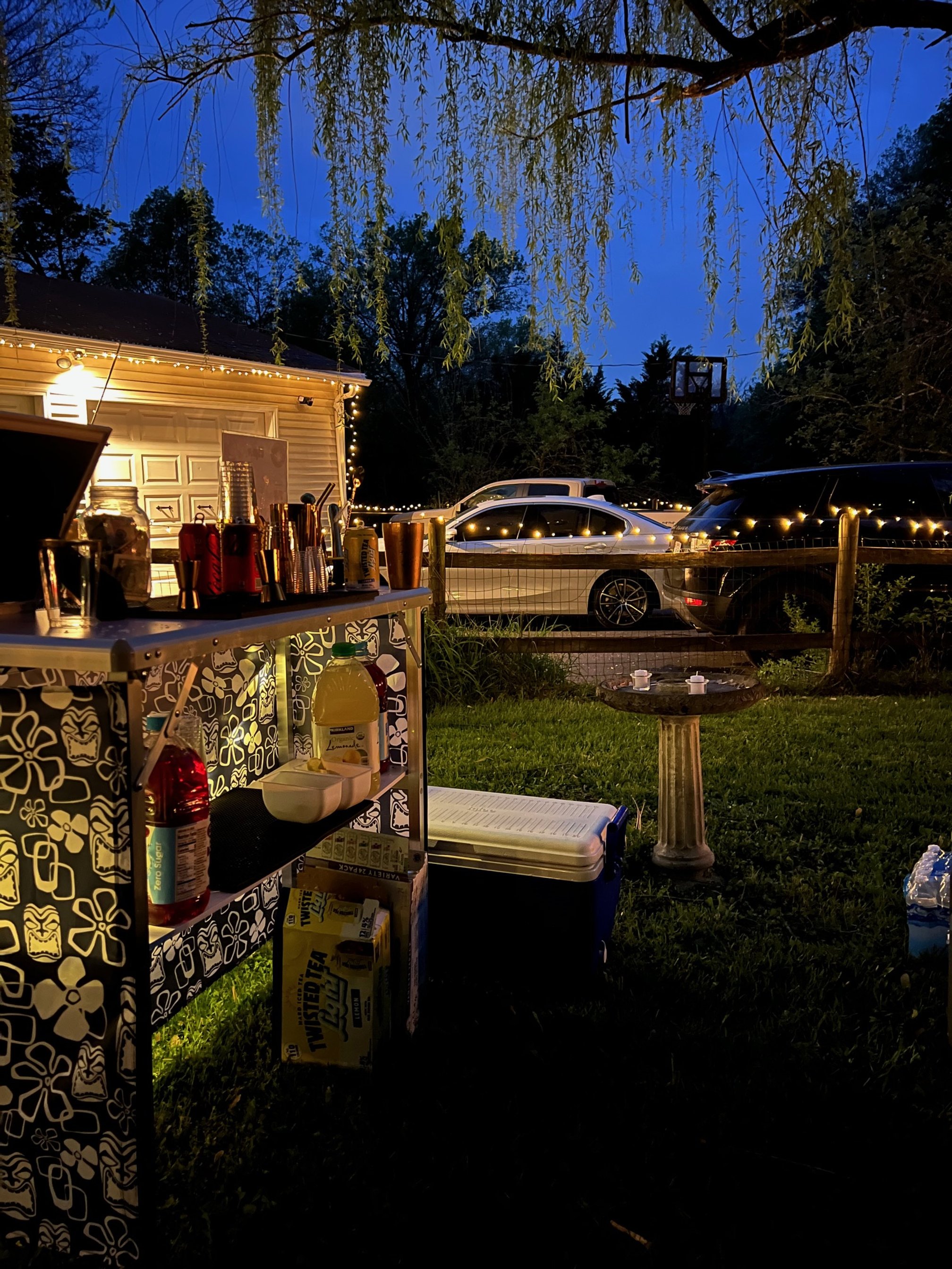 Outdoor evening scene with a bar setup including bottles and mixers on a patterned table. String lights illuminate the area with cars parked and a house in the background. Grass, trees, and a birdbath are visible.
