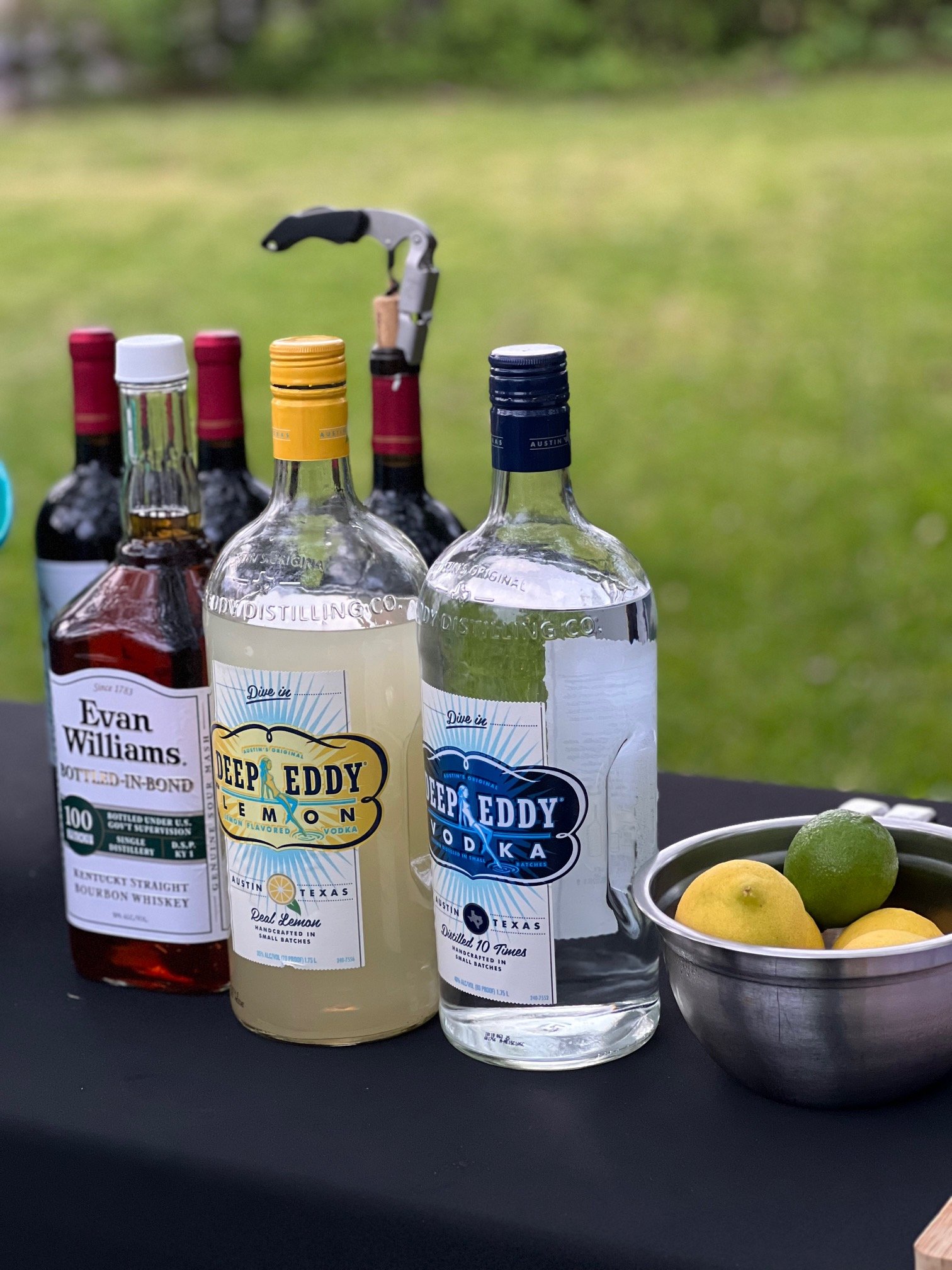An outdoor setup featuring a bottle of bourbon labeled Evan Williams, a bottle of Deep Eddy Lemon vodka, and a bottle of Deep Eddy Vodka. Next to them is a metal bowl containing lemons and a lime. A corkscrew and additional bottles of wine are visibl