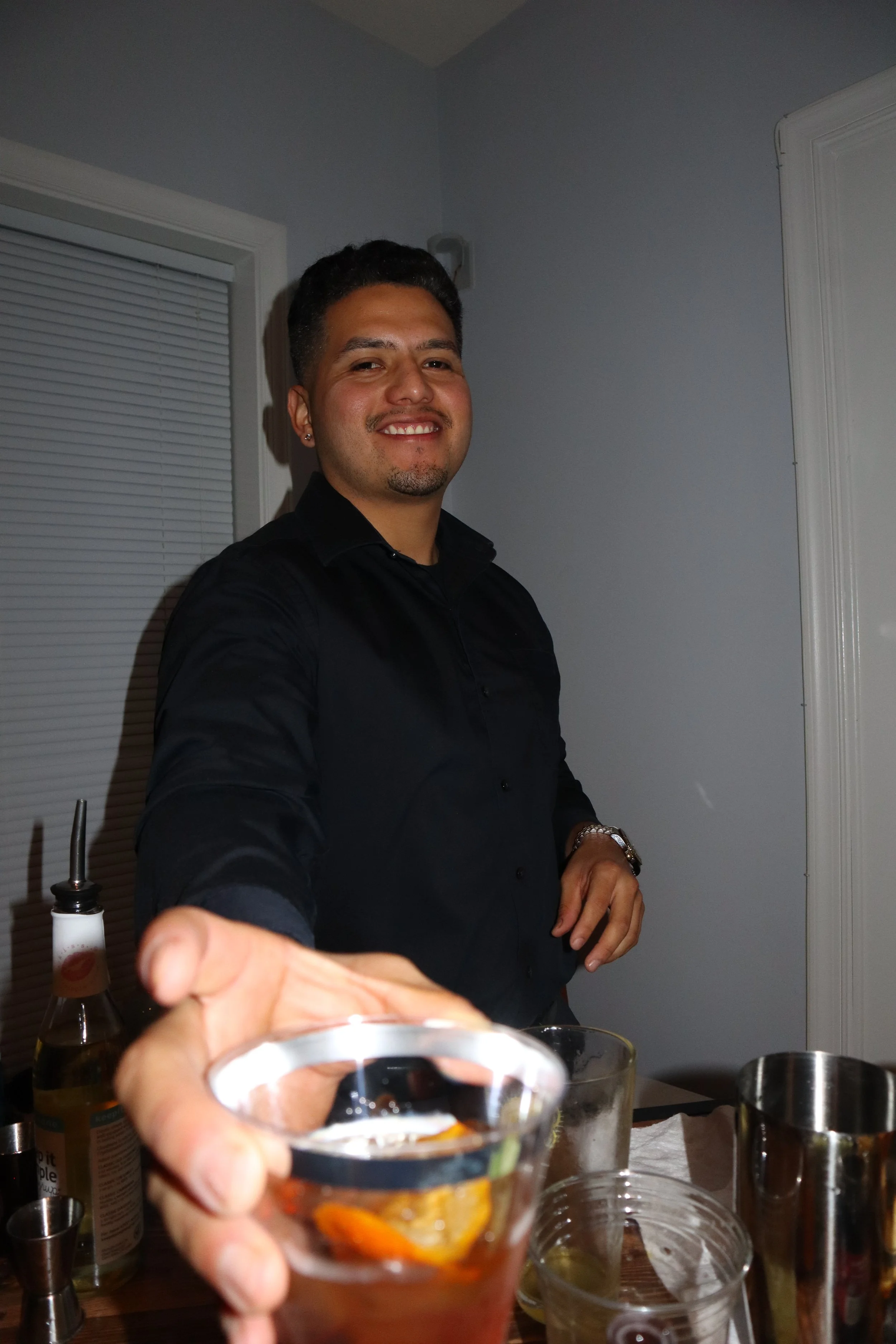 Person in black shirt smiling and handing a drink in a clear plastic cup, standing by a table with drink items.