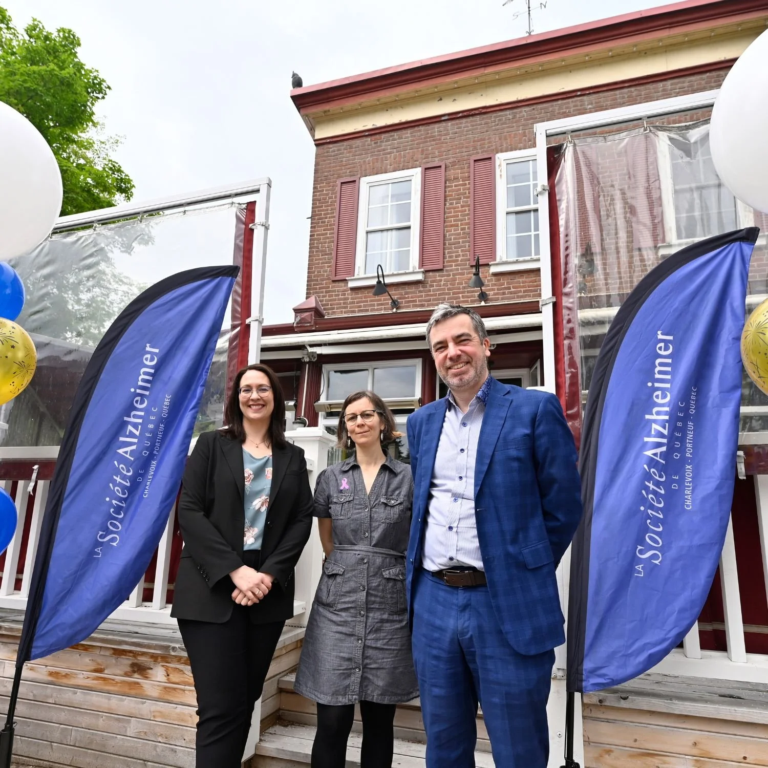 Trois personnes debout devant une maison en briques, avec des drapeaux bleus portant l'inscription 'La Société Alzéimer'.