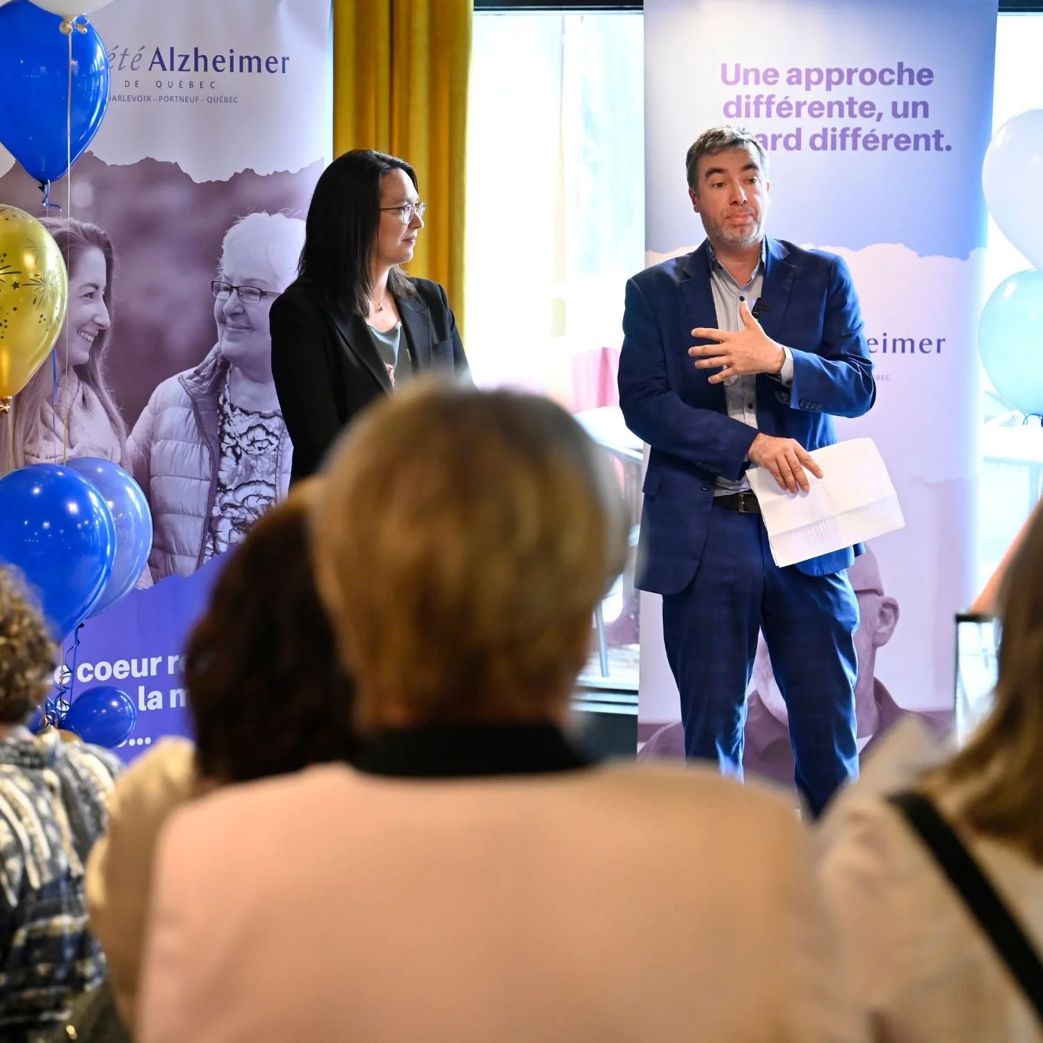 Une conférence ou réunion sur la maladie d'Alzheimer avec trois intervenants, un homme en costume bleu qui parle et une femme à gauche qui écoute, dans une salle décorée avec des ballons bleus et des affiches sur Alzheimer