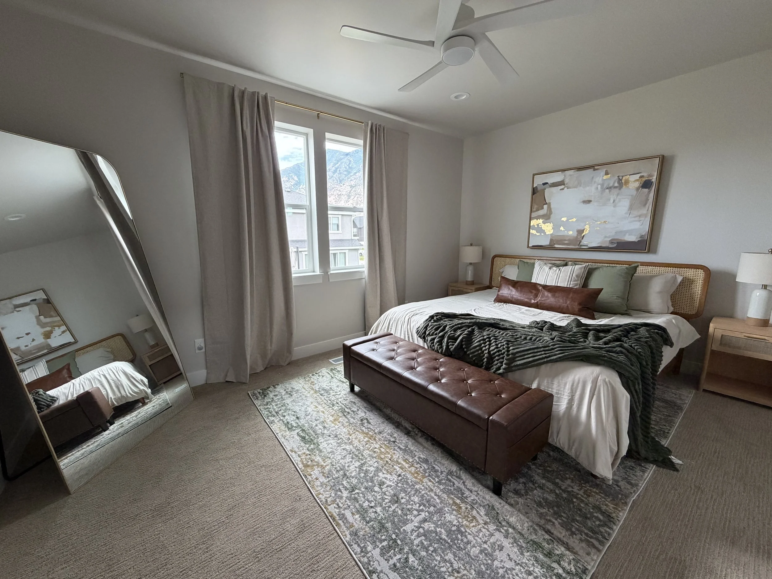 A bedroom with a bed, pillows, a nightstand with a lamp, a large framed abstract painting on the wall, a window with curtains showing mountains outside, a ceiling fan, and a mirror reflecting part of the room.