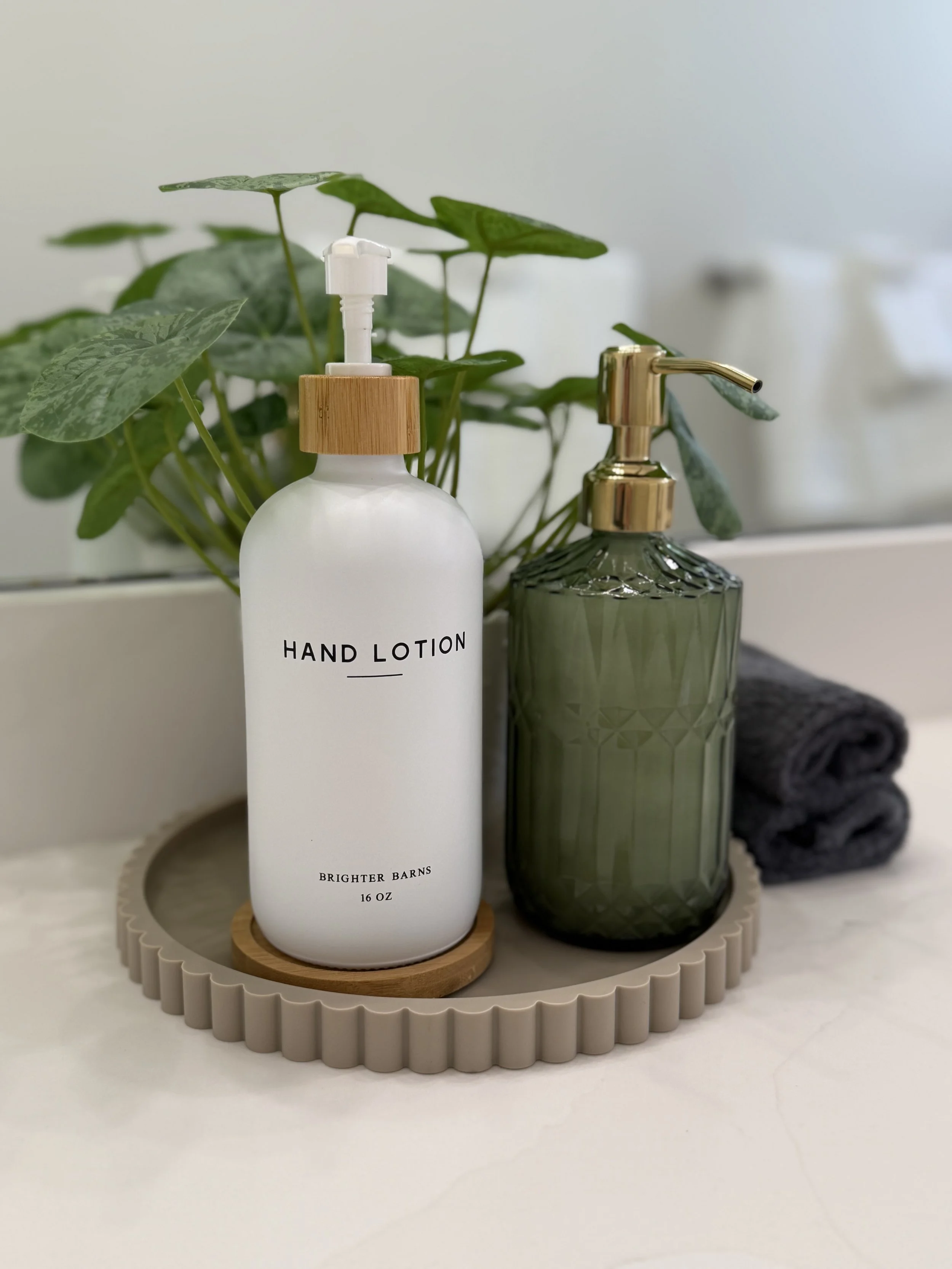 A bathroom countertop with a white hand lotion bottle labeled 'Brighter Barns' on a round wooden tray, next to a green textured soap dispenser with a gold pump, a faucet, a black rolled towel, and a plant with green leaves.