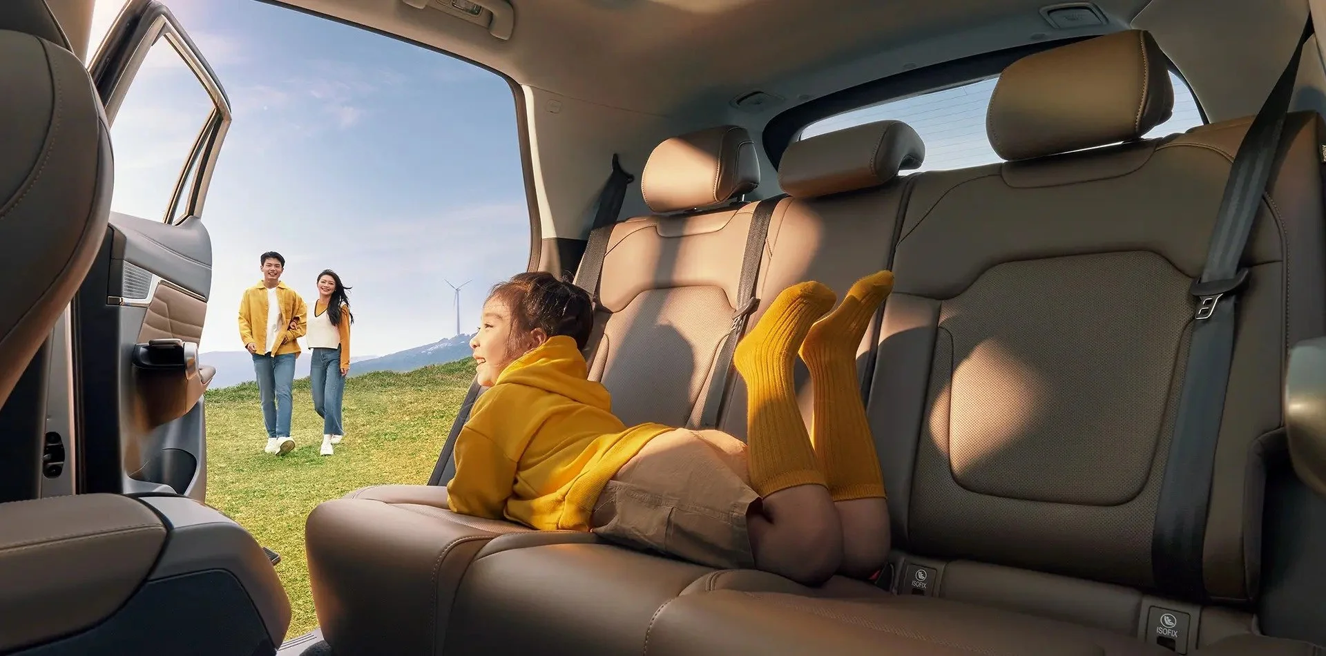 Mujer acostada en el asiento trasero de un vehículo, mirando hacia afuera, con dos personas caminando en el campo, árboles, montaña y molino de viento en el fondo.