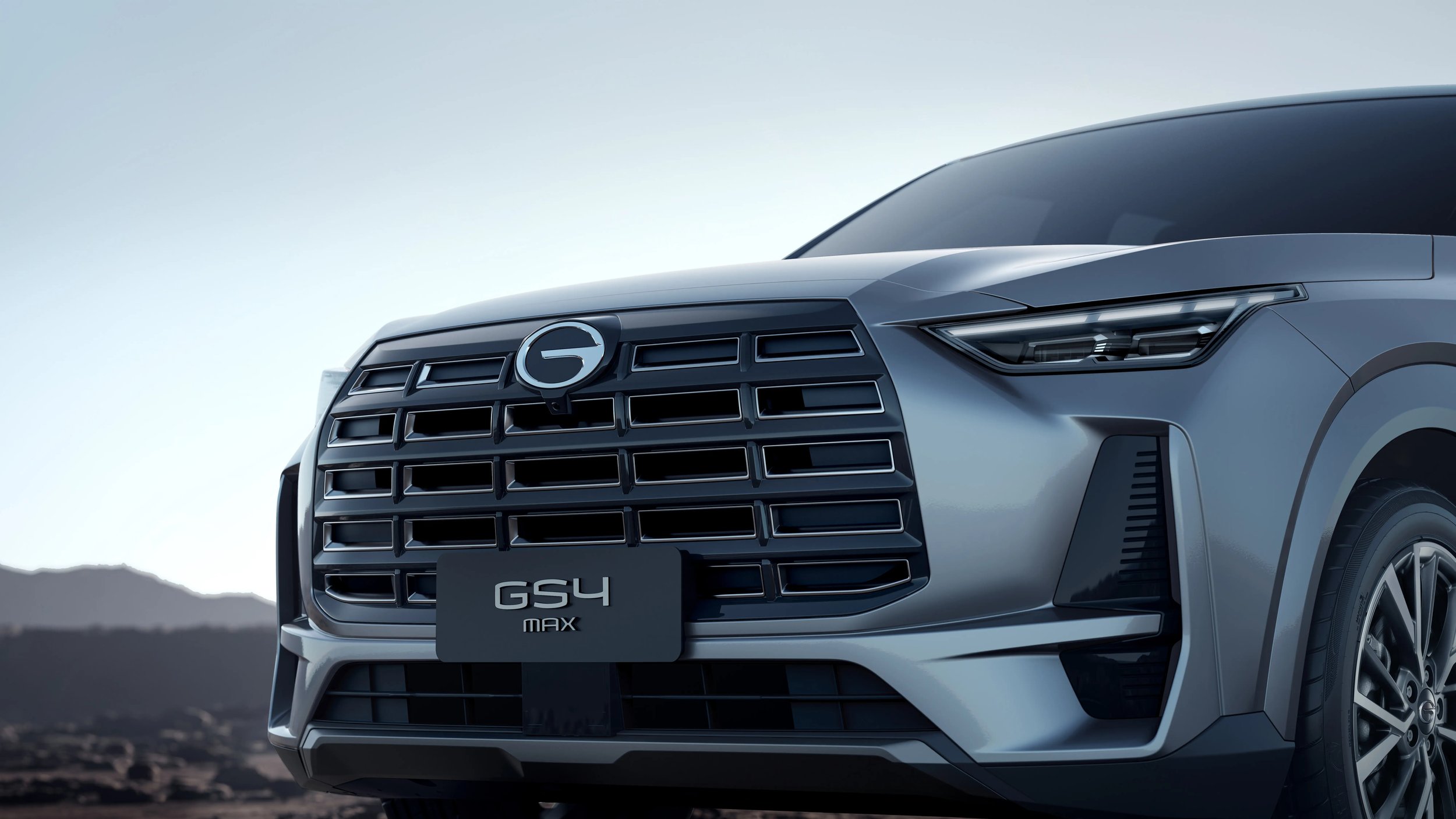 Frontal de un carro gris con parrilla grande, en un paisaje montañoso al atardecer o amanecer.