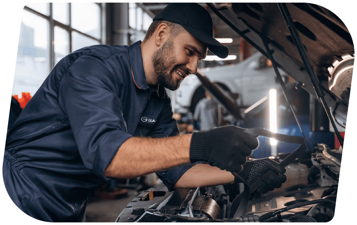 Un mecánico sonriente trabaja en la parte del motor de un coche en un taller de autos, usando guantes negros y una gorra negra.