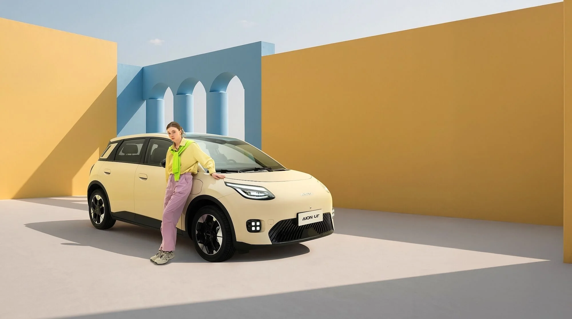 Una mujer posa junto a un automóvil de diseño futurista en un fondo colorido con paredes azules y amarillas, y arcos.