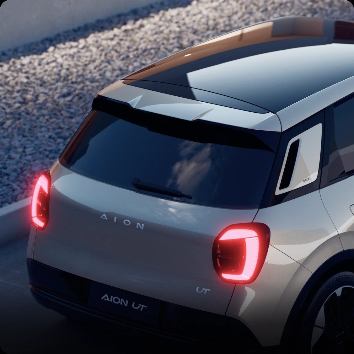 Parte trasera de un coche eléctrico blanco, marca AION, modelo UT, con luces traseras encendidas, aparcado junto a un muro con piedra y una superficie de grava.