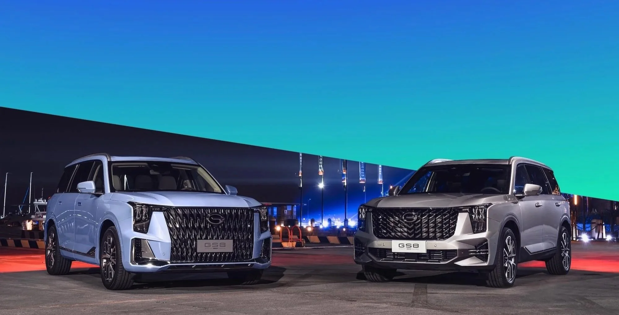 Dos autos SUVs plateados aparcados en un puerto con fondo de cielo al atardecer y banderas, uno a la izquierda y otro a la derecha, con diseño moderno y parrillas grandes.