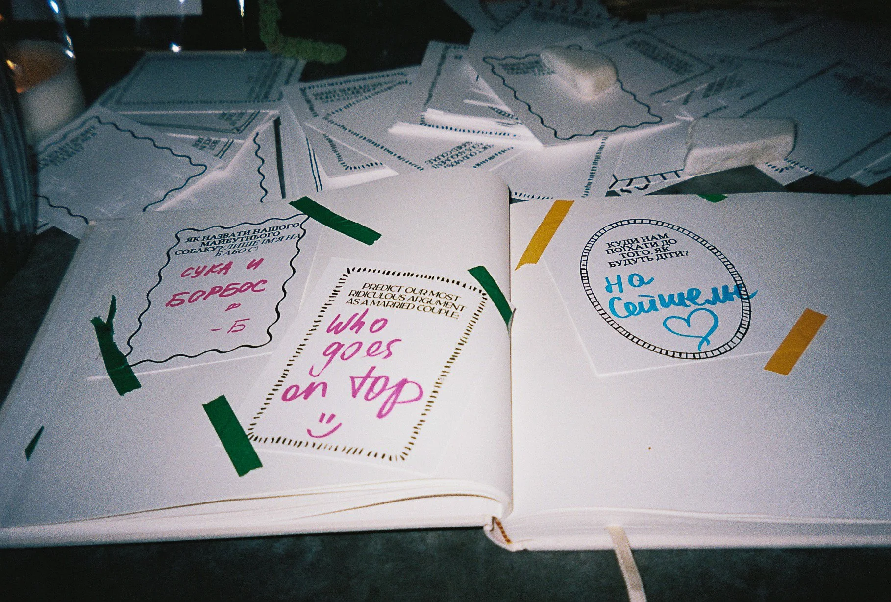 Open book with various handwritten notes and quotes on pages, surrounded by scattered papers. Notable note says 'Who goes on top' with a smiley face, and another says 'Аксенель', with some writing in Ukrainian.