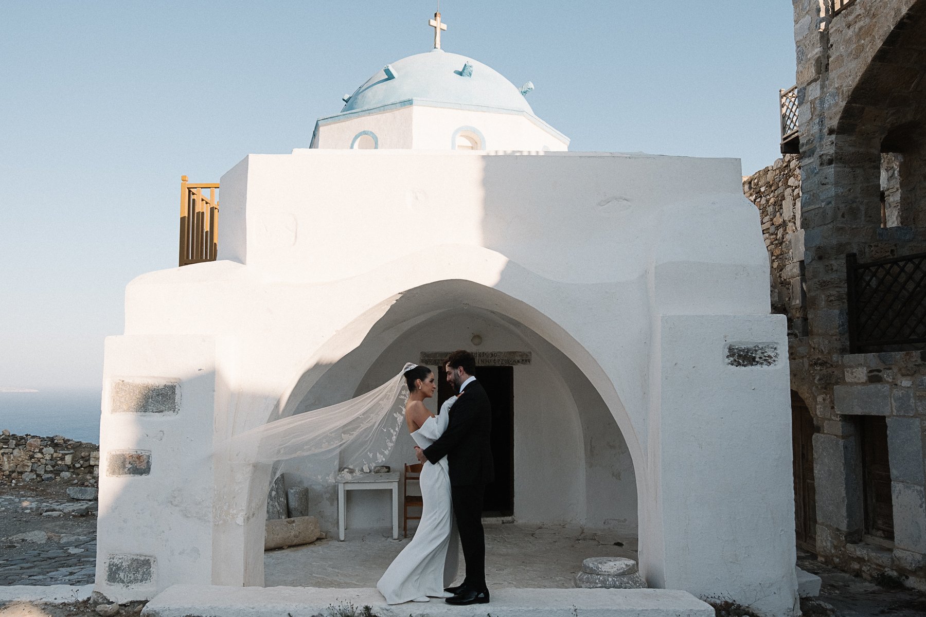 Bride and groom in wedding attire standing close together outside a white chapel with a blue dome and a cross on top, on a sunny day.
