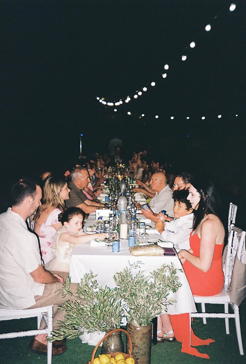 A large dinner party with many guests seated along a long table decorated with candles and flowers, under a string of hanging lights at night.