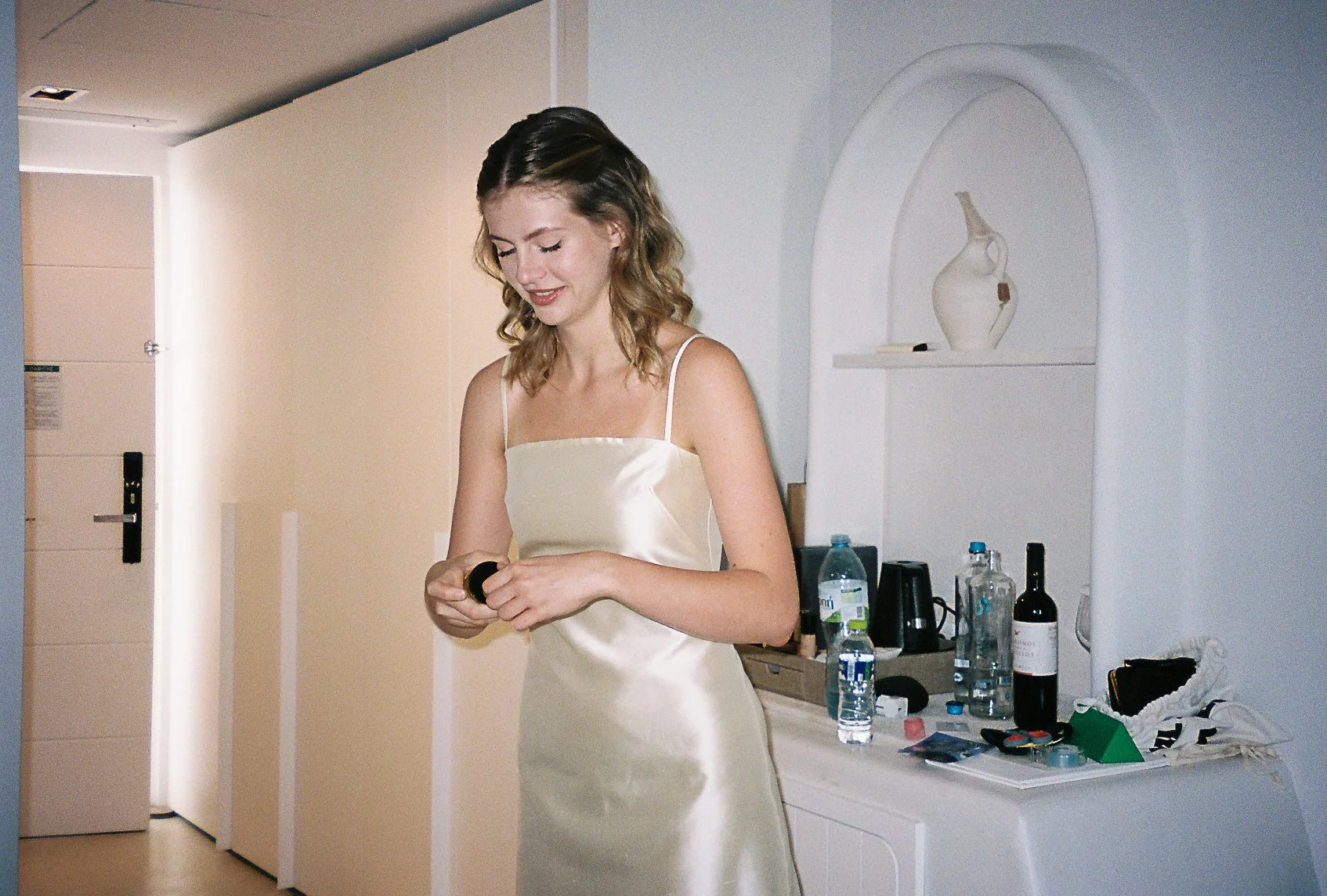 A young woman in a satin slip dress standing inside a room with a white dresser and bottles on top, smiling as she looks at a small object in her hands.