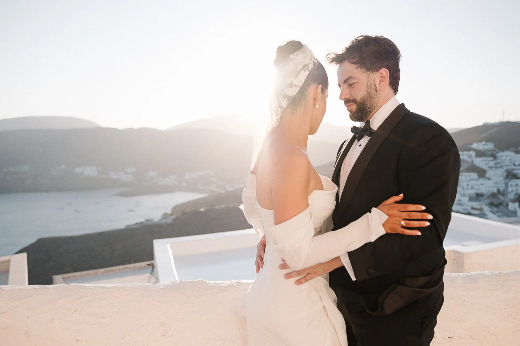 A newlywed couple in wedding attire sharing a close moment outdoors with a scenic coastal background, during sunset.