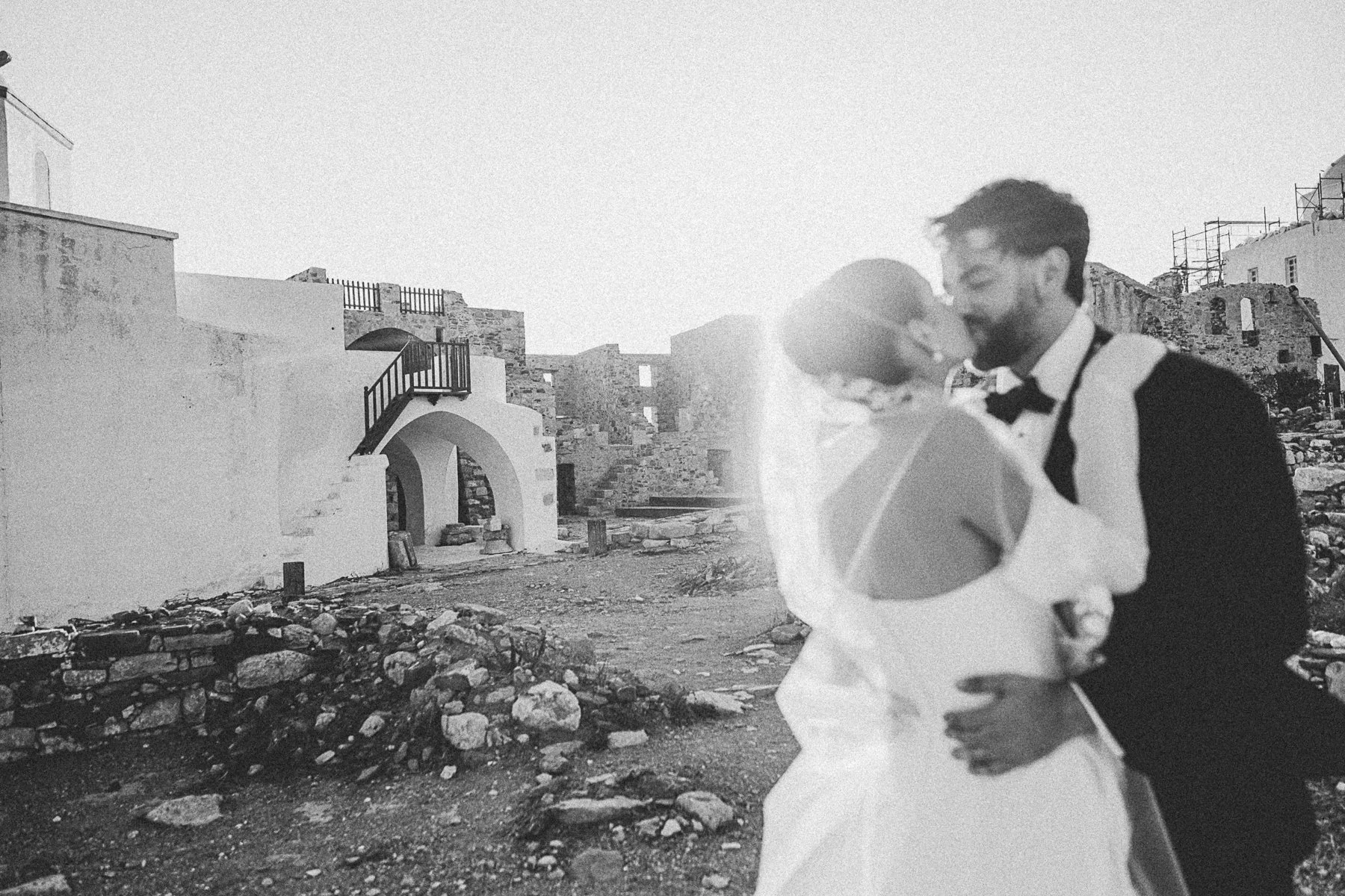 A black and white photo of a bride and groom dancing outdoors with a rustic stone building in the background.
