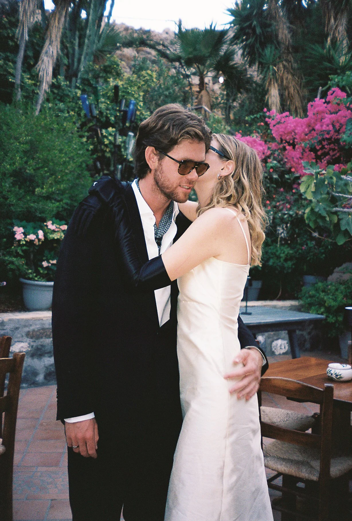 A man and woman embracing outdoors during sunset, surrounded by trees and pink flowers, with some chairs and a table nearby.
