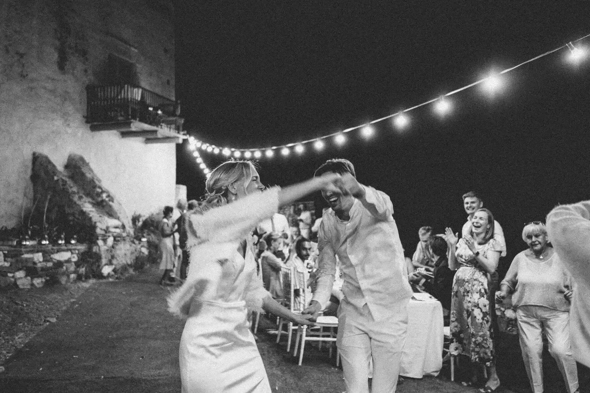 A wedding reception outdoor at night with string lights, people dancing and celebrating, a bride and groom are dancing and smiling, surrounded by happy guests.