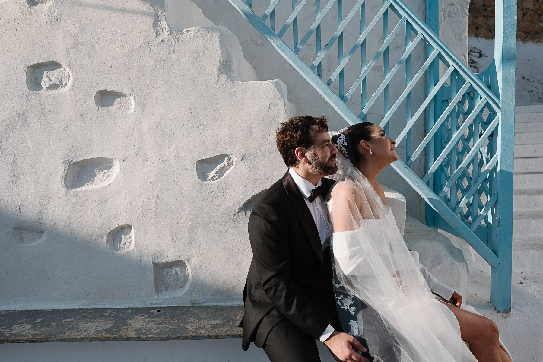 A bride and groom sitting against a white stucco wall with blue stairs in the background, smiling and enjoying a moment together.