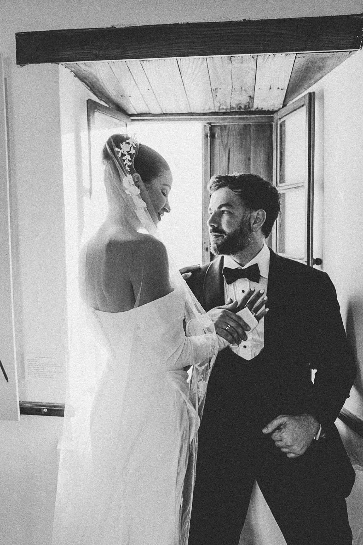 A black and white photo of a bride and groom inside a room near a window. The bride wears a wedding dress with an off-shoulder design, and the groom is in a tuxedo with a bow tie. They are looking at each other affectionately, with the groom holding 