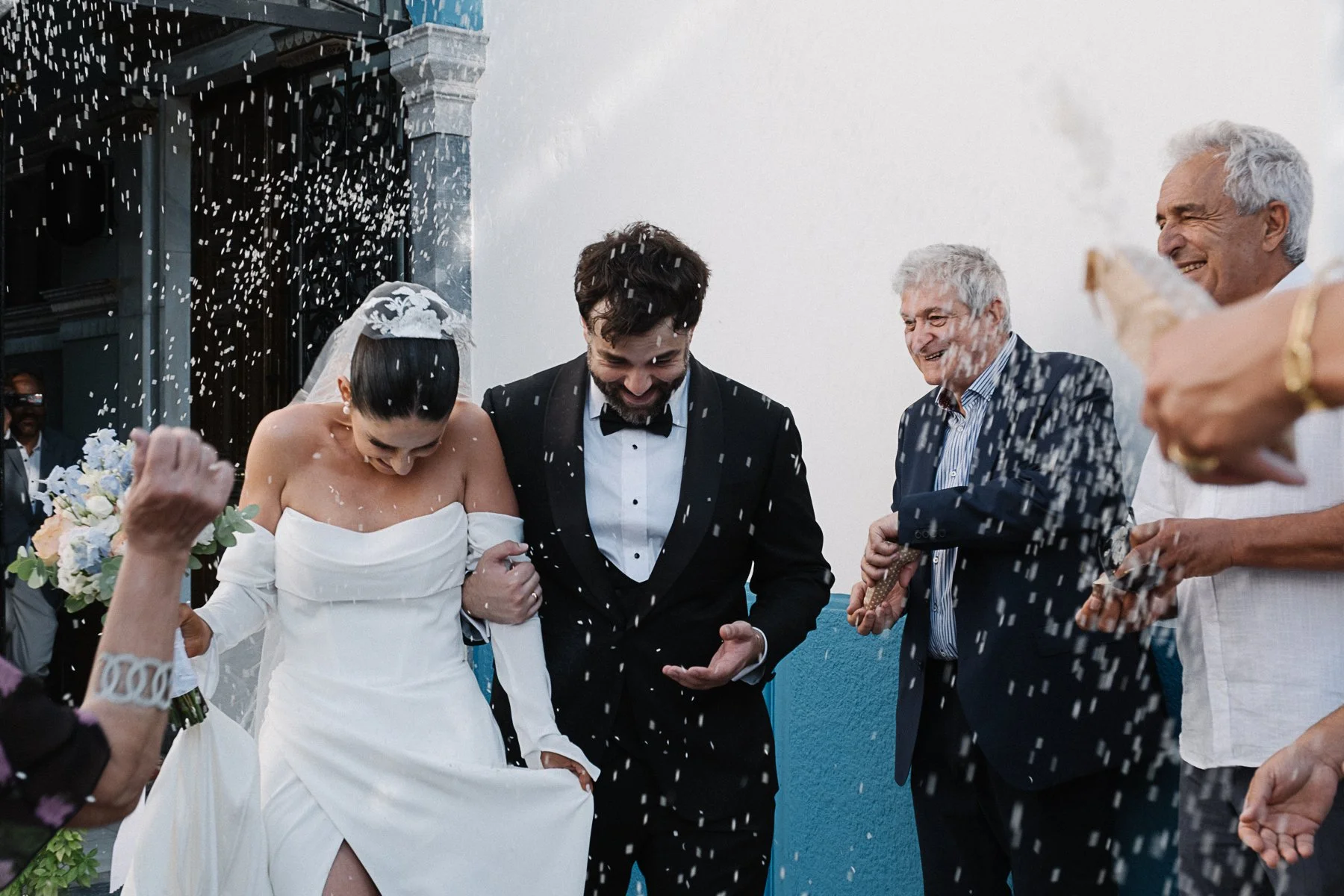 People celebrating a wedding with confetti outside a building, including a bride in a white dress and veil, and a groom in a black tuxedo, smiling and holding hands.