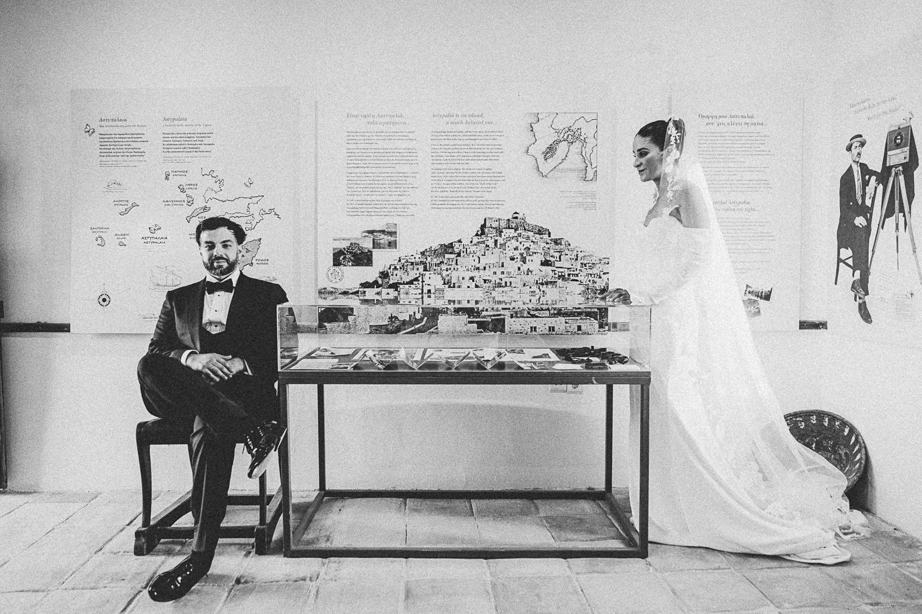 A man in a tuxedo sitting on a chair with crossed legs and a woman in a wedding dress standing beside a display case at an exhibit with maps and posters of Greece on the wall behind them.