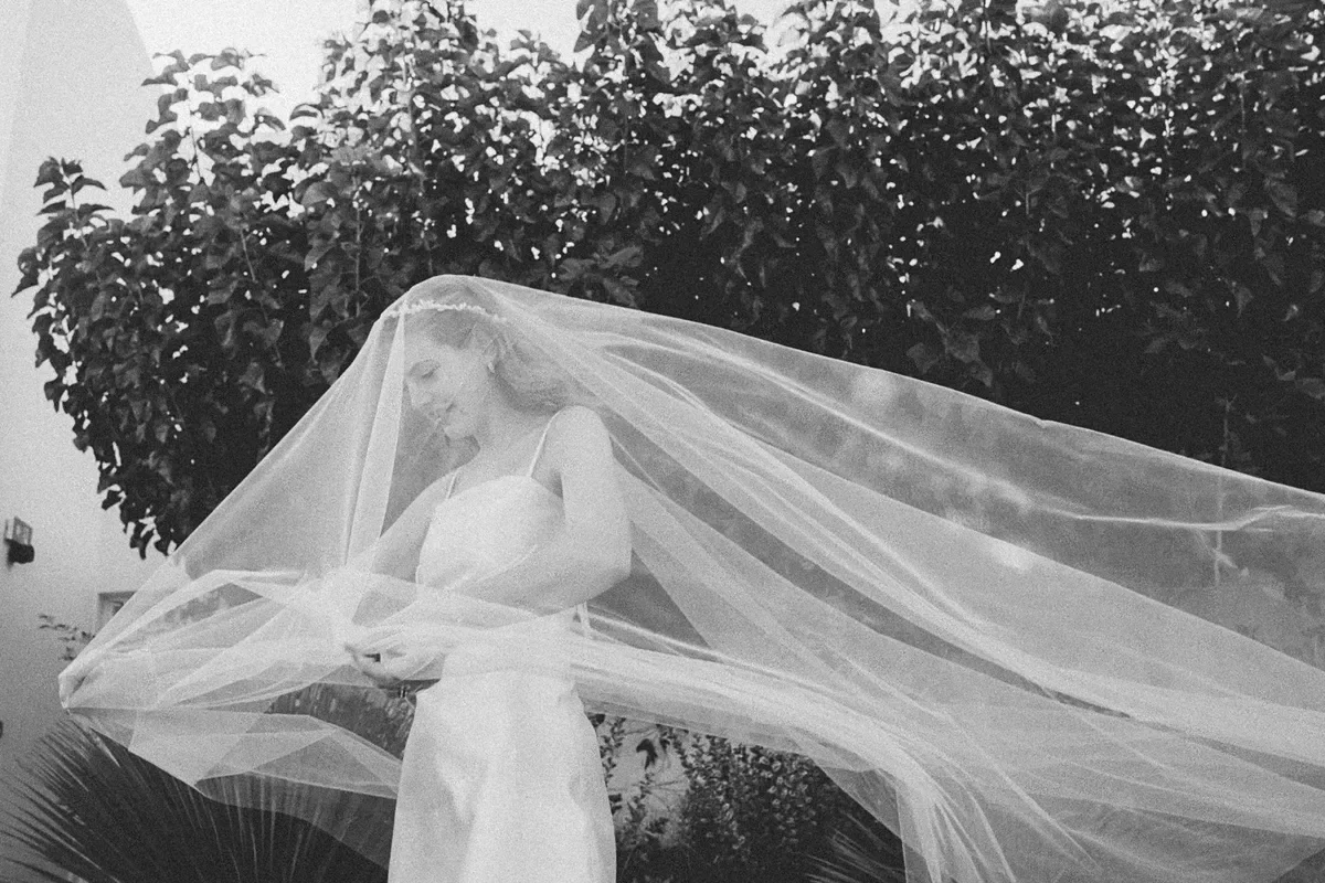 A bride in a wedding dress with a long veil standing outdoors, with trees in the background.