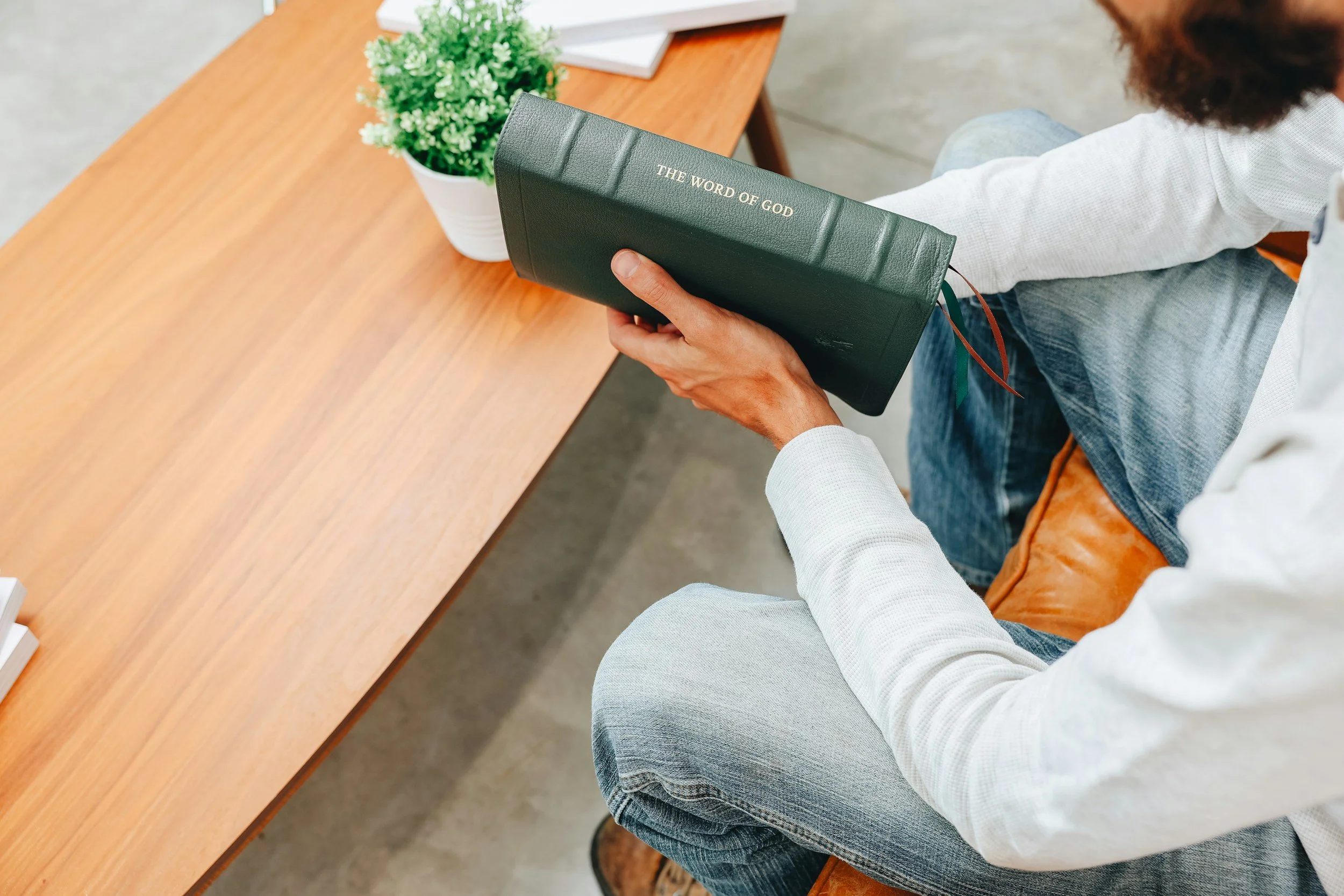 A man sitting at a wooden table reading a large, dark green Bible with gold lettering that says 'The Word of God' on the cover. There is a white pot with a green plant on the table.