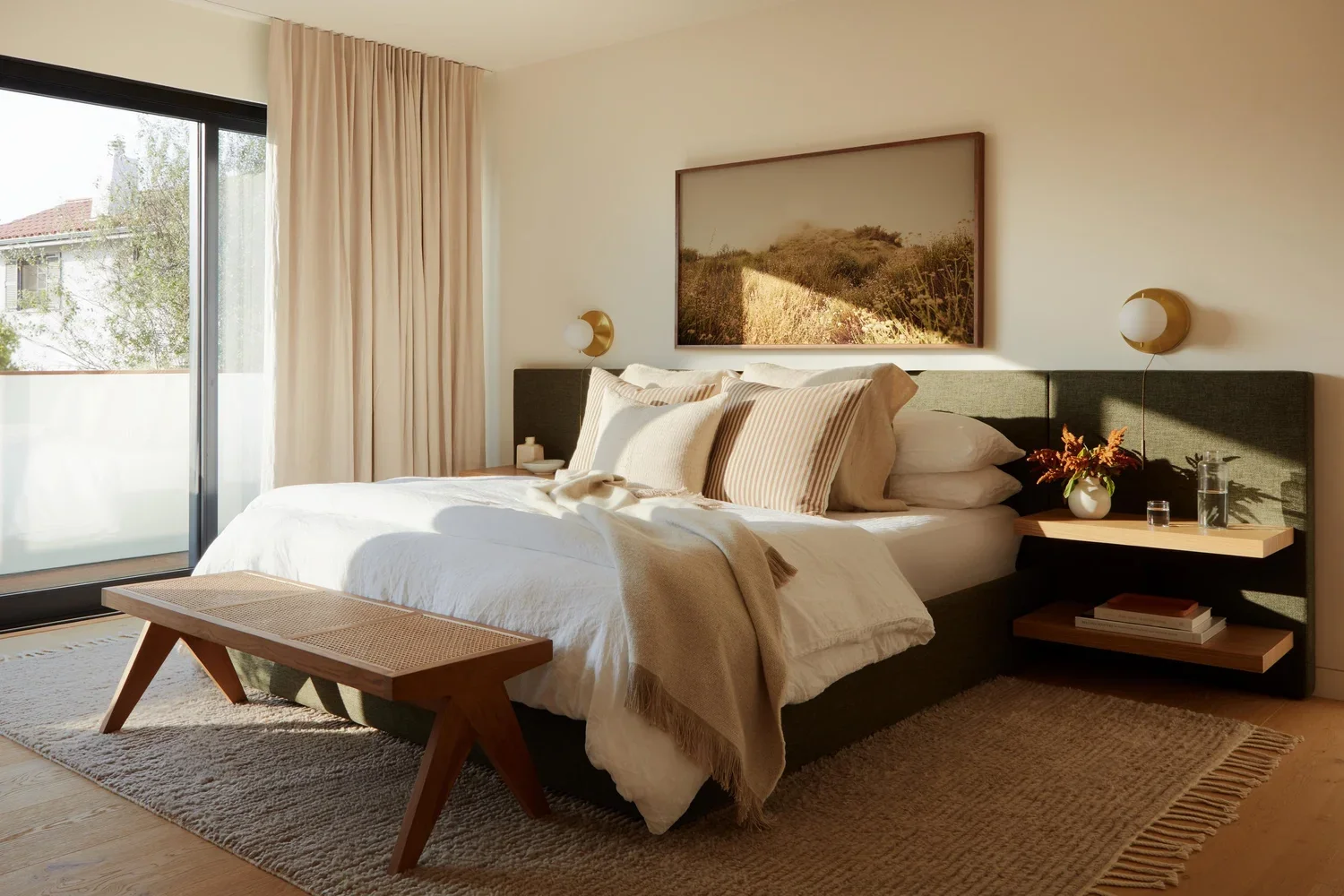 Studio Lifestyle–designed bedroom in a Santa Monica Canyon home featuring a neutral upholstered platform bed, floating wood nightstands, brass globe sconces, woven bench, and floor-to-ceiling linen drapery with warm natural light.