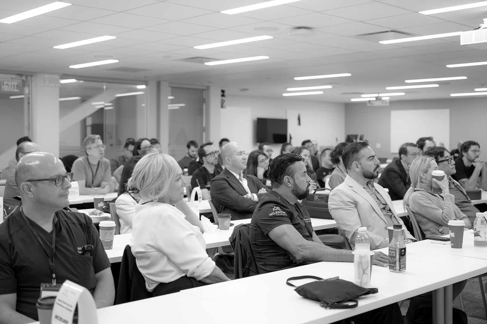 A group of people attending a conference or seminar in a large, well-lit room, sitting at tables and listening attentively.