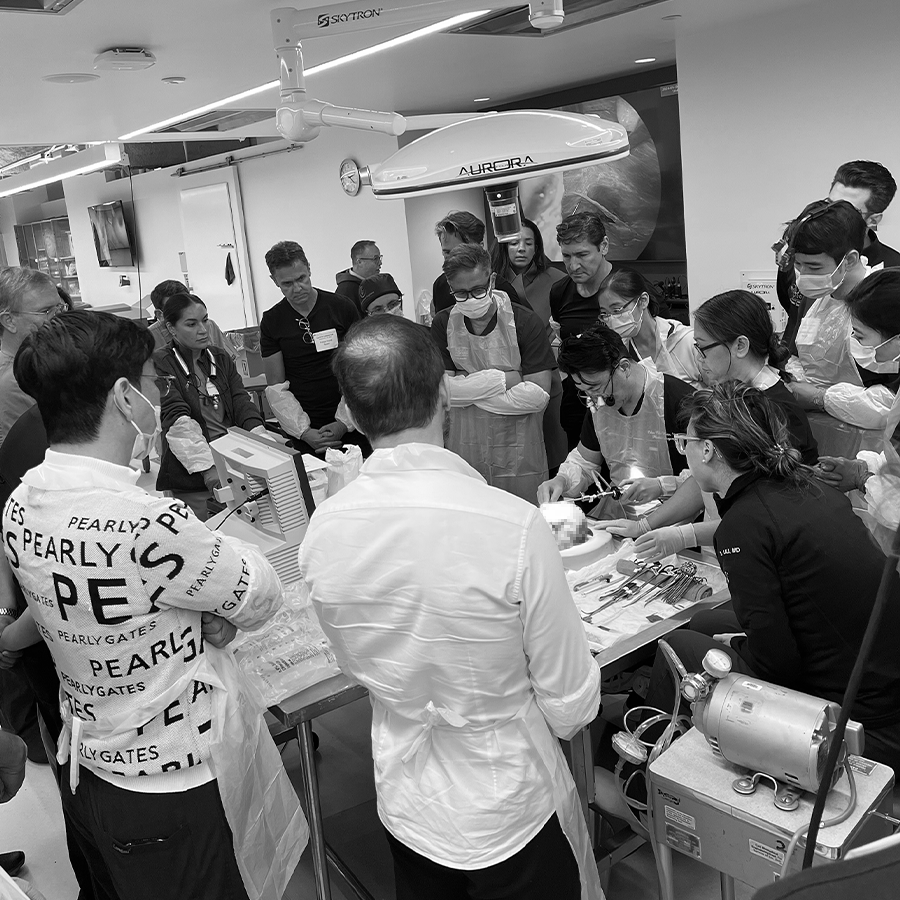 Group of medical professionals and students gathered around a medical table during a training or demonstration in a clinical setting, with surgical instruments and equipment present.