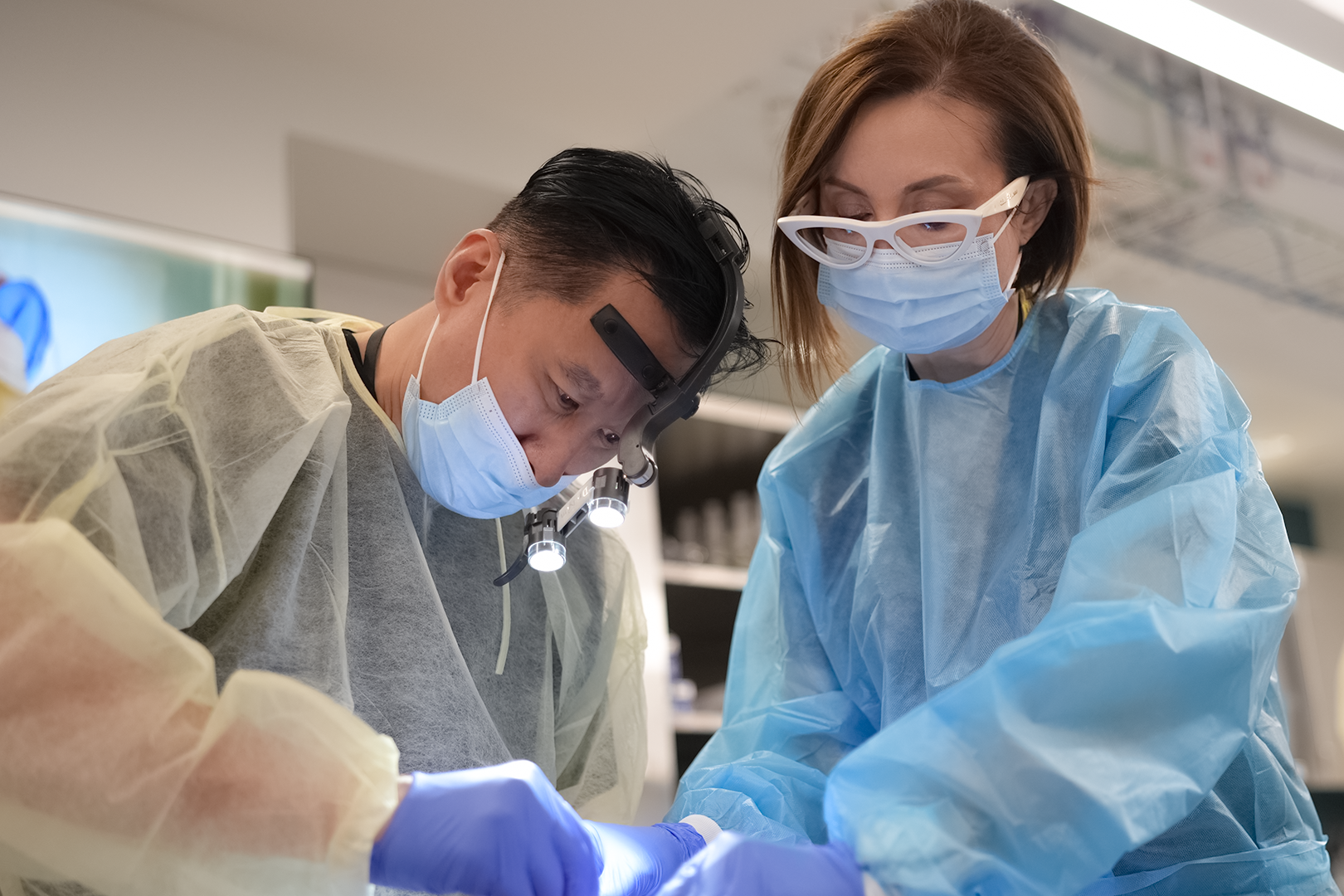 Two medical professionals, a male surgeon and a female assistant, performing a surgical procedure in an operating room, wearing masks, gowns, and protective eyewear.