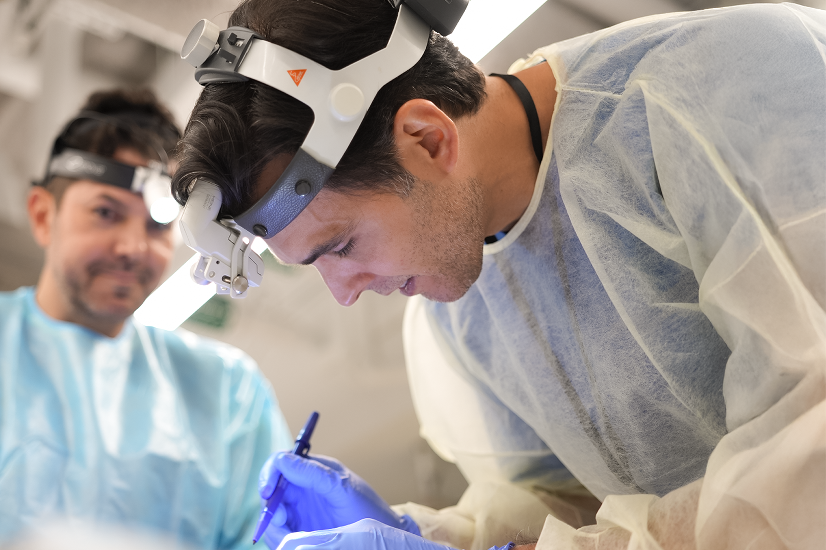 Two male surgeons wearing magnifying headlamps and surgical gowns performing surgery, one is focused on the surgical site, the other is observing.