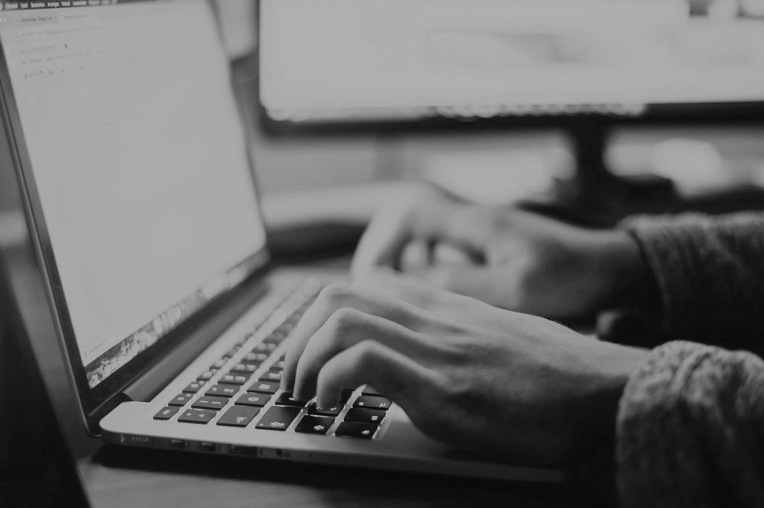 Person typing on a laptop keyboard with multiple computer screens in the background, in black and white.