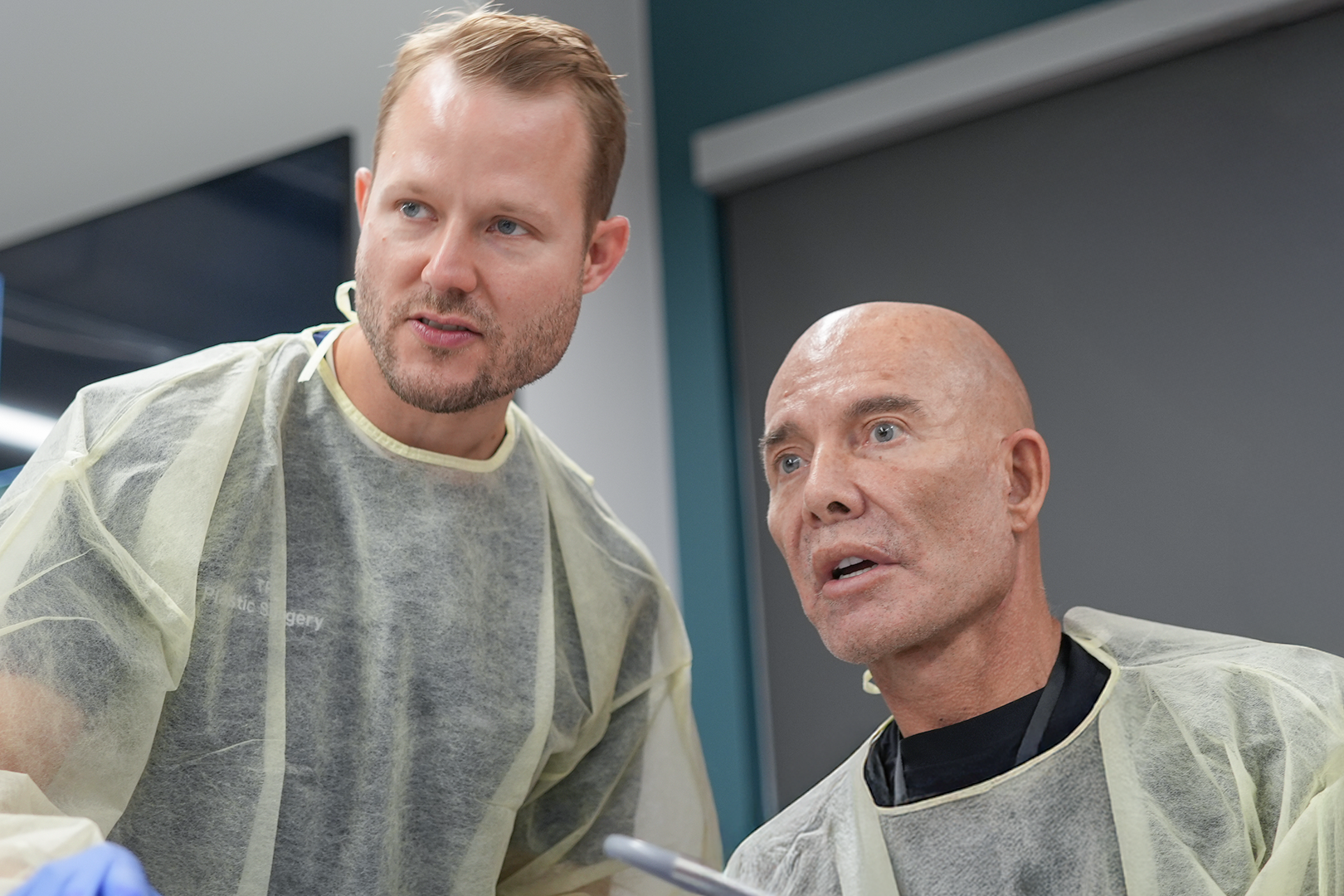 Two men in scrubs, one standing and one sitting, engaged in a medical discussion in a clinical setting.