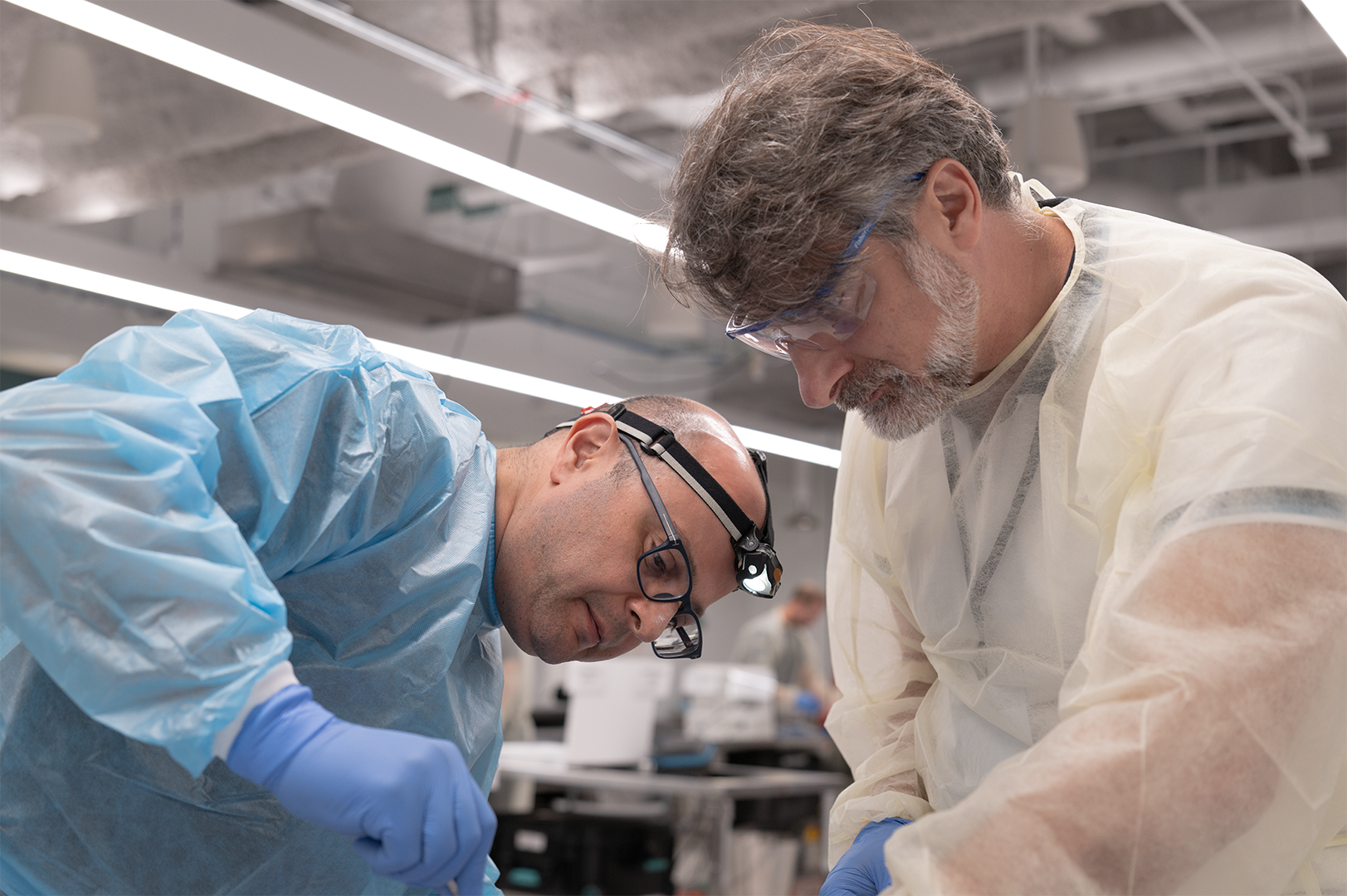 Two surgeons working closely, wearing protective clothing, gloves, and safety glasses, focused on a task.