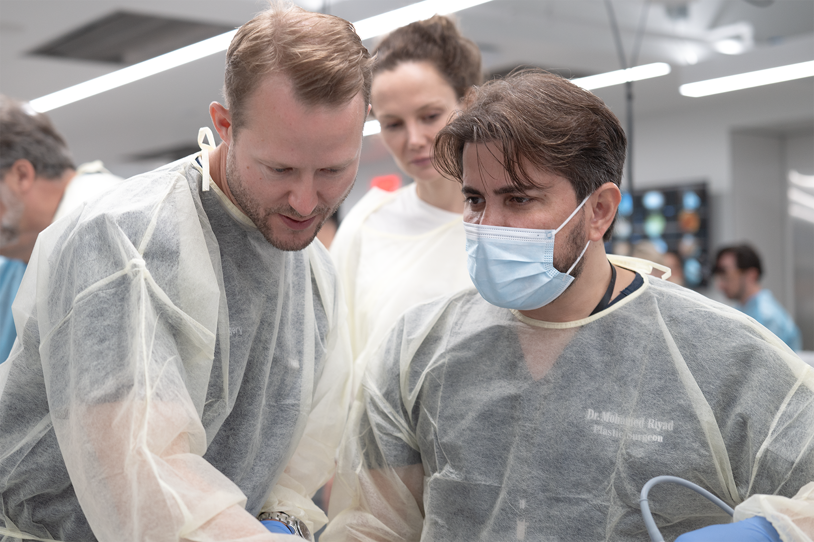 Two medical professionals in protective gowns and masks perform a procedure, with a woman observing in the background, in a clinical setting.