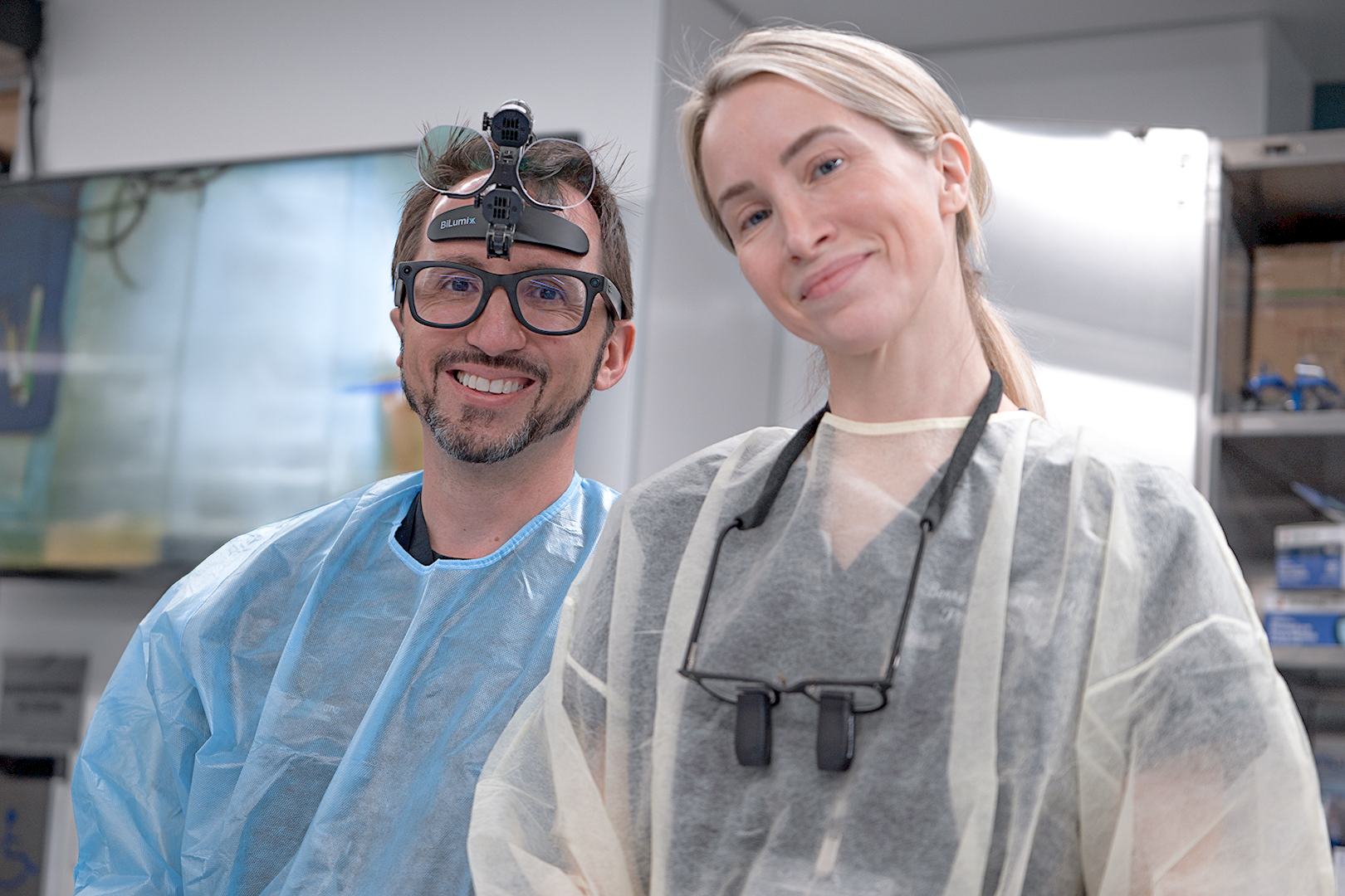 Two medical professionals, a man and a woman, smiling at the camera in a clinical setting, both wearing scrubs and gloves, with medical equipment and shelves in the background.