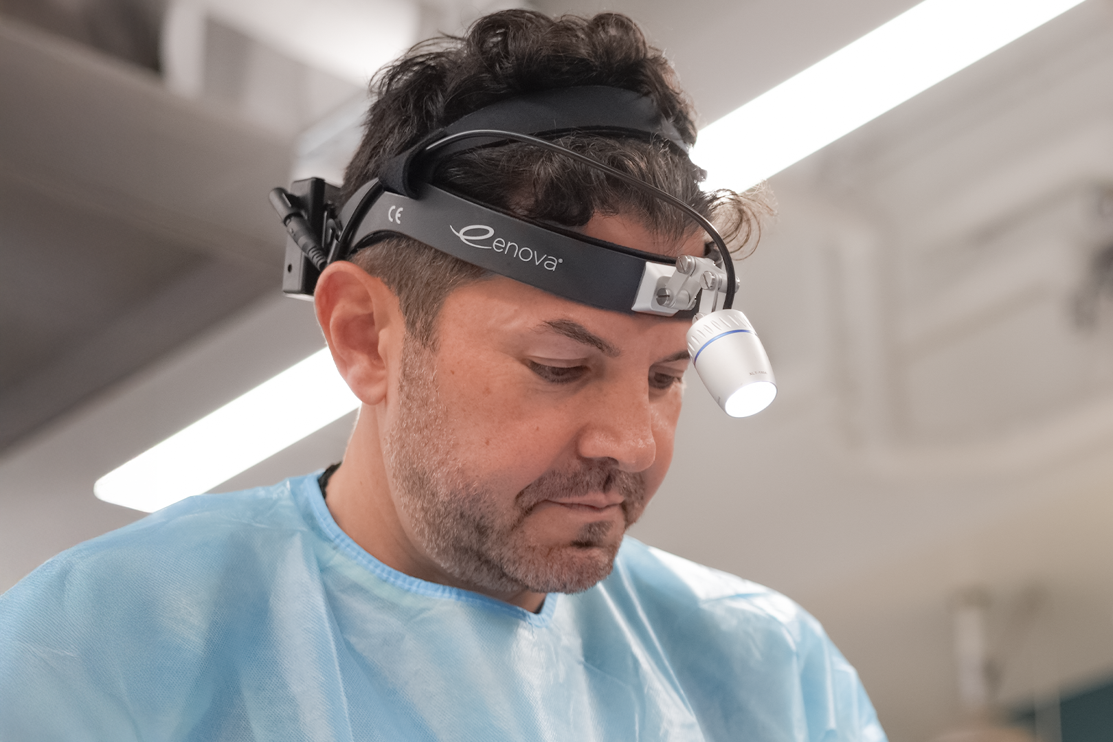 A male doctor wearing a headlamp and light blue scrubs, working in a clinical setting.