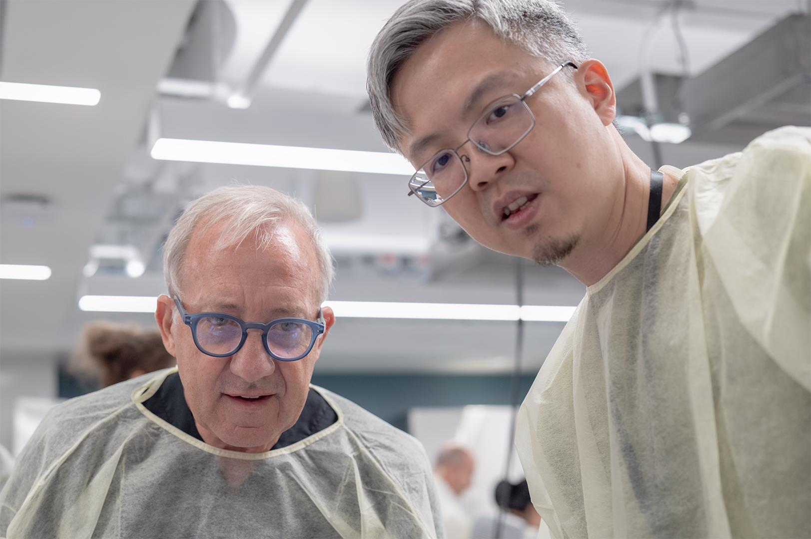 Two men wearing protective gowns and glasses in a laboratory or medical setting, closely examining something.