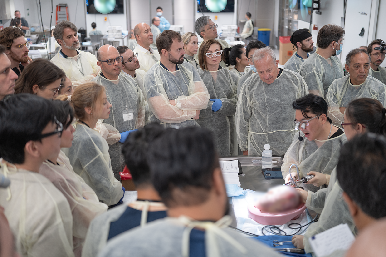 Medical professionals in a lab gathered around a surgical procedure, some wearing masks, gloves, and protective gowns, with a person performing a demonstration.