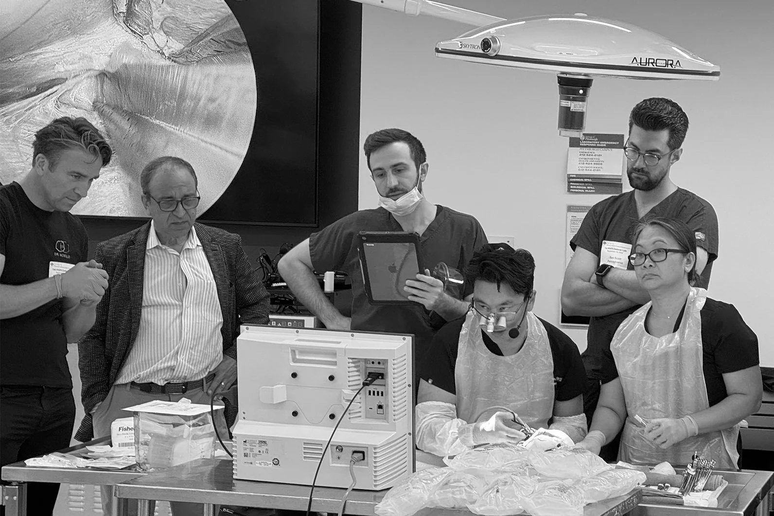 A group of medical professionals and a visitor observing a surgical procedure in a hospital or medical training setting, with some wearing gloves, aprons, and surgical glasses, and one person using a surgical microscope.