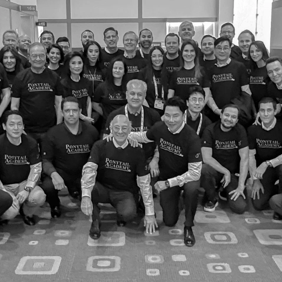 A large group of people wearing matching Ponytail Academy t-shirts posing together indoors, some kneeling in front and others standing behind, smiling for the camera.
