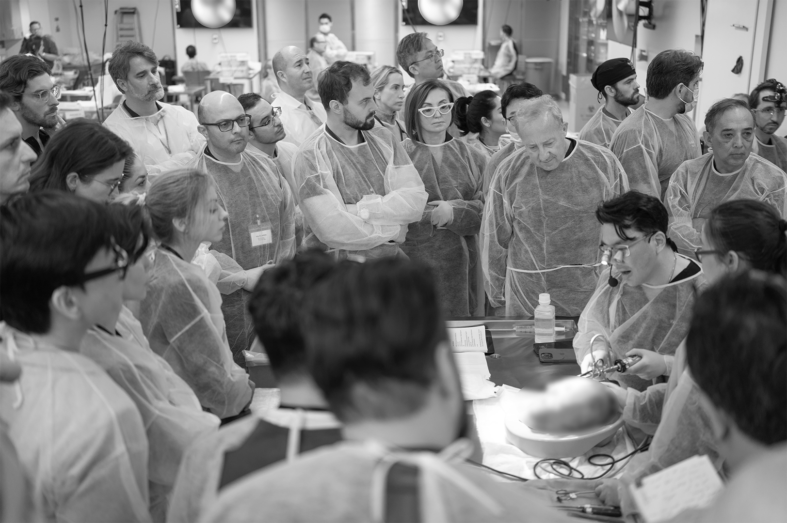 Group of medical professionals and students gathered around a patient in a hospital or clinical setting, with some wearing masks and examination gowns, focused on a procedure.