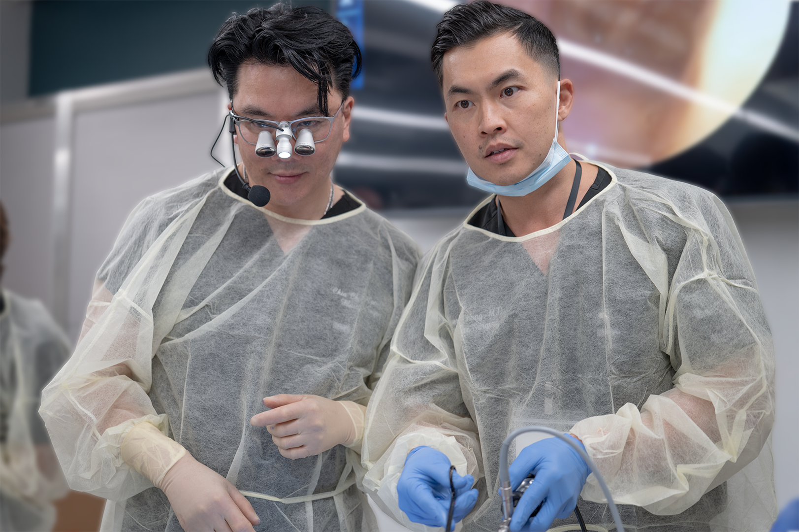 Two male surgeons in operating room wearing protective gowns, gloves, and masks. One surgeon is using a surgical instrument, while the other is observing. Both are focused on a procedure.