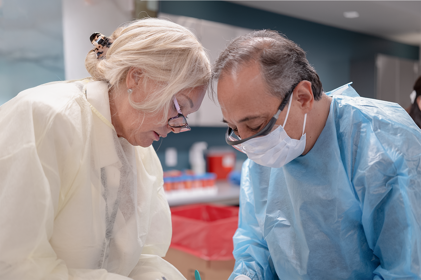 Two medical professionals wearing masks and protective gowns working closely together in a clinical setting.