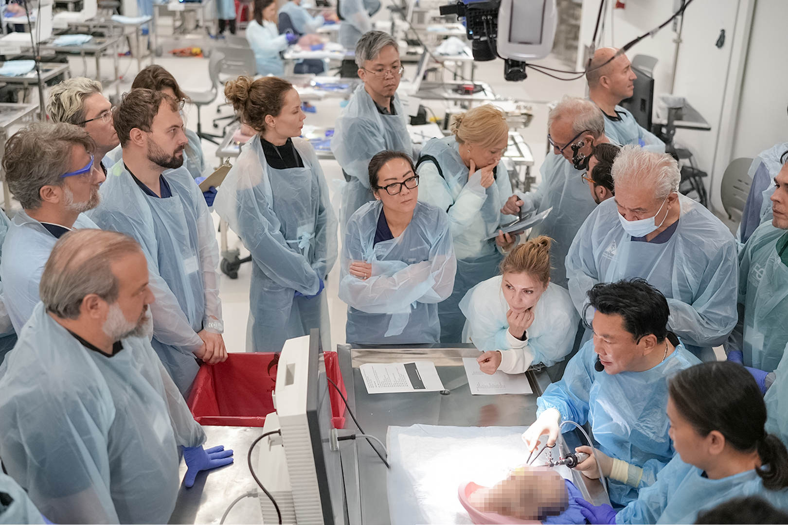 A group of medical professionals and researchers in surgical scrubs gathered around an operating table, observing a procedure involving a baby. Some are taking notes, others are closely watching the procedure, with a bright surgical light illuminatin
